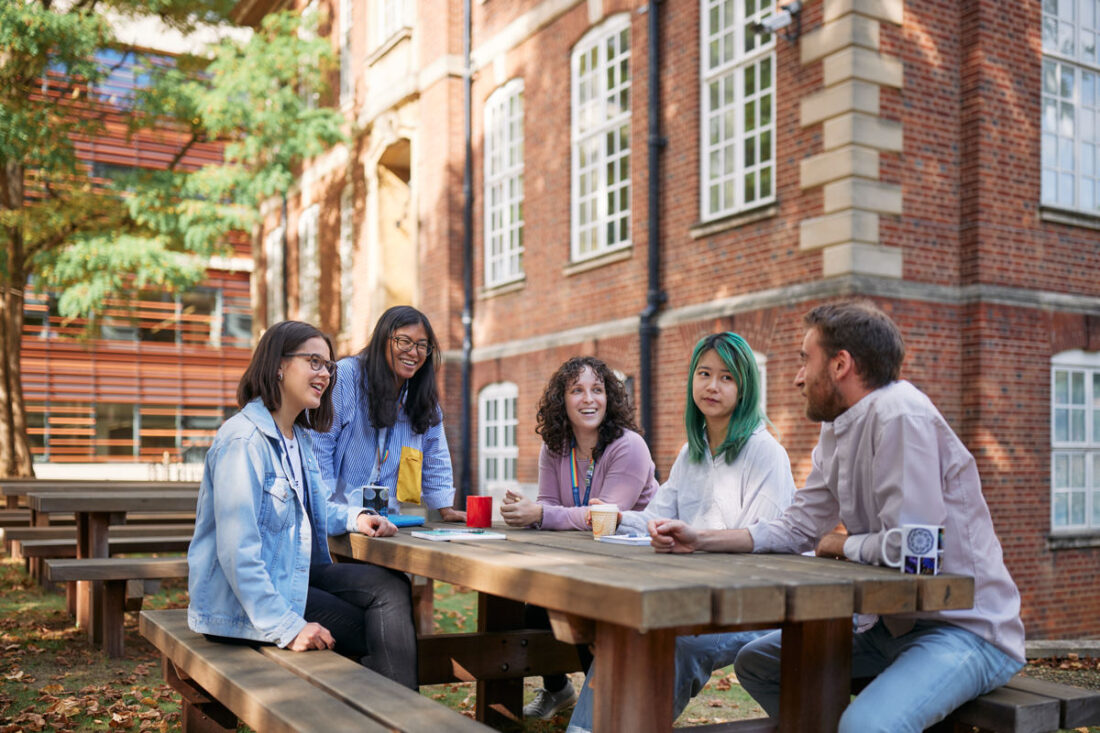 group of people sit around a table outside the Dunn School, laughing and chatting