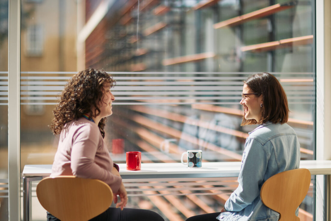 2 people talk to each other while having a hot drink, close to a window facing the OMPI building